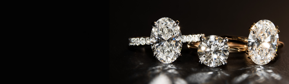 Three diamond rings displayed on a dark reflective surface, including an oval-cut diamond in white gold setting with round sidestones, a round brilliant-cut diamond in a yellow gold solitaire setting, and another oval-cut diamond in a yellow gold solitaire setting.