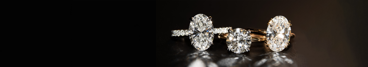 Three diamond rings displayed on a dark reflective surface, including an oval-cut diamond in white gold setting with round sidestones, a round brilliant-cut diamond in a yellow gold solitaire setting, and another oval-cut diamond in a yellow gold solitaire setting.