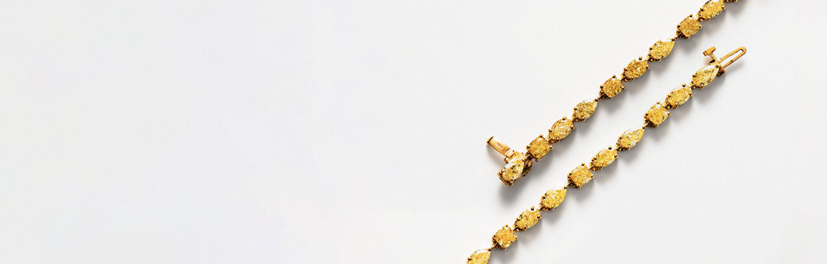 A close-up of a gold tennis bracelet with an alternating layout of pear, cushion and oval yellow diamonds, laid diagonally on a clean white background.
